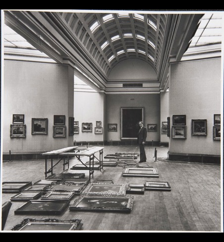 Sir Philip Hendy, Director, standing in Room 29. He is surrounded by paintings laid out on the floor in preparation for a rehang of the room.