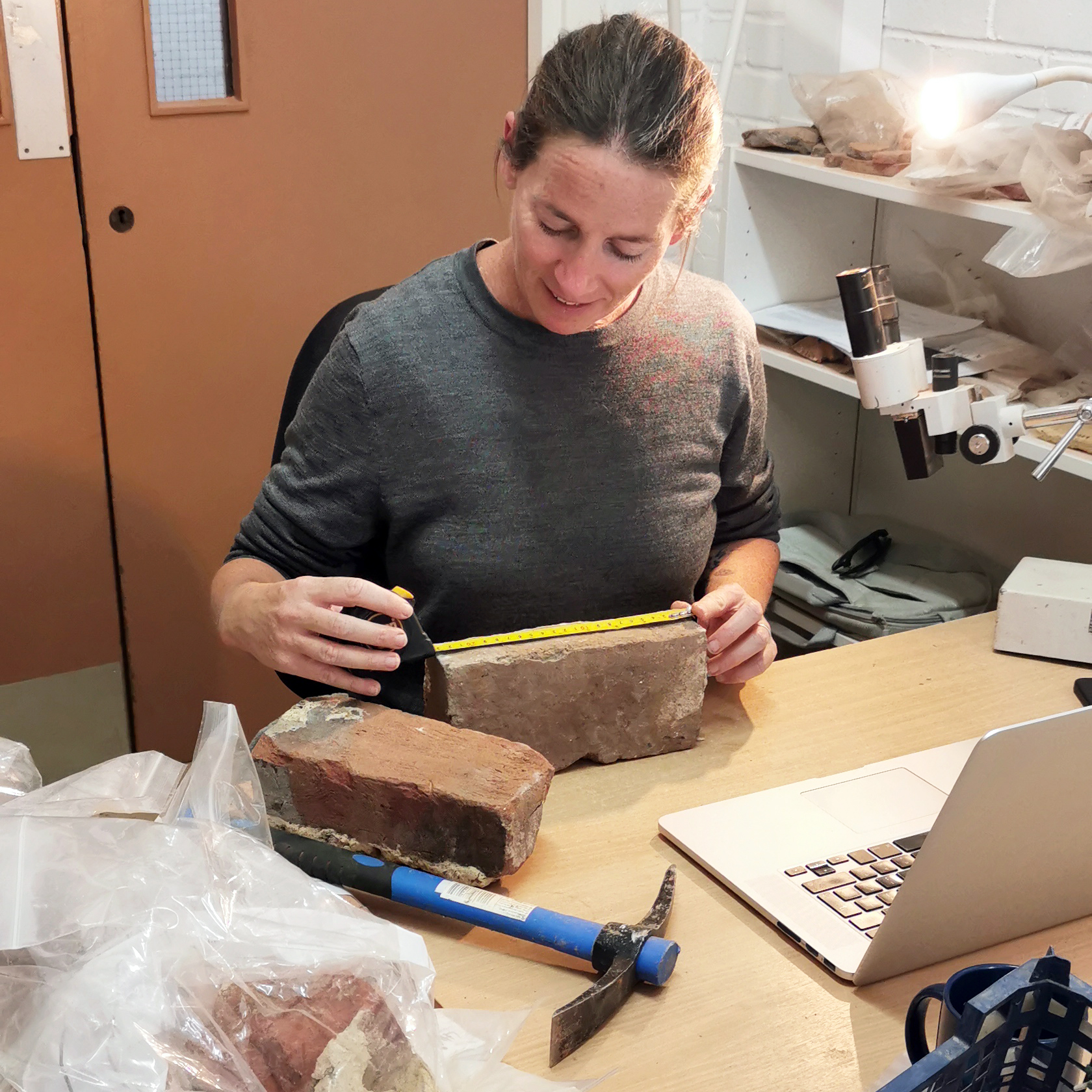 an archaeologist in a lab measures a brick with a yellow tape measure. Around her on the desk are other bricks, a small pickaxe for removing brick fragments, and a microscope.