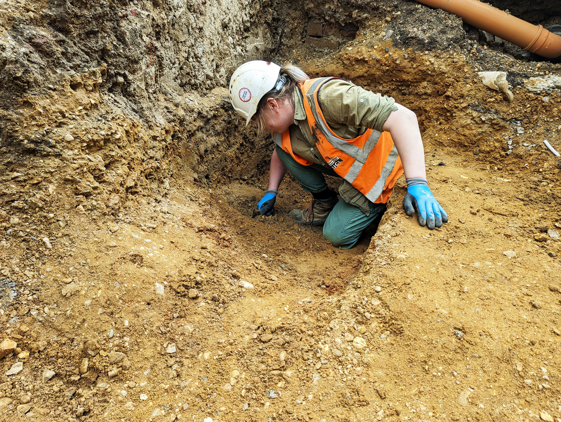 An archaeologist crouches in an excavated ditch, using a trowel to scrape at the soil in front of them