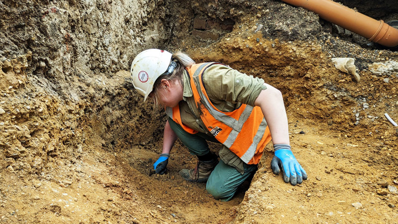 An archaeologist crouches in an excavated ditch, using a trowel to scrape at the soil in front of them