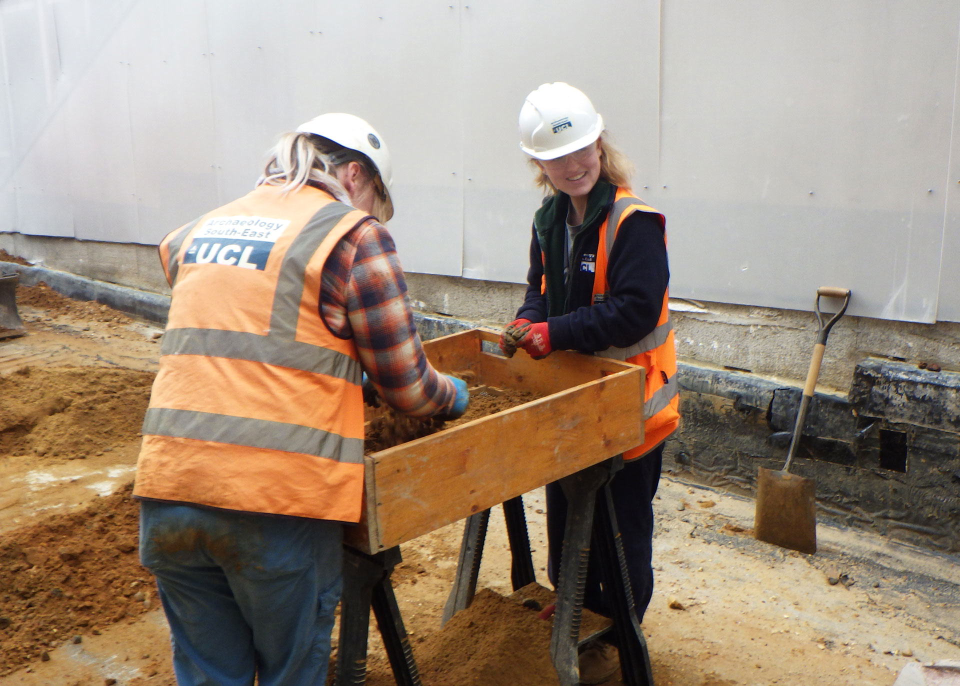 Two archaeologists stand over a large wooden box with a mesh base sieving artefacts from soil. One smiles towards the camera