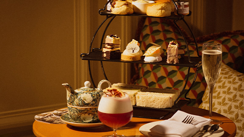 A tiered stand of finger sandwiches and pastries. In the foreground is a festive red cocktail, glass of champagne and decorative tea pot