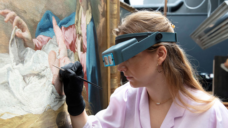 A conservator wearing magnifying glasses holds a fine paintbrush to a panel by Peruzzi