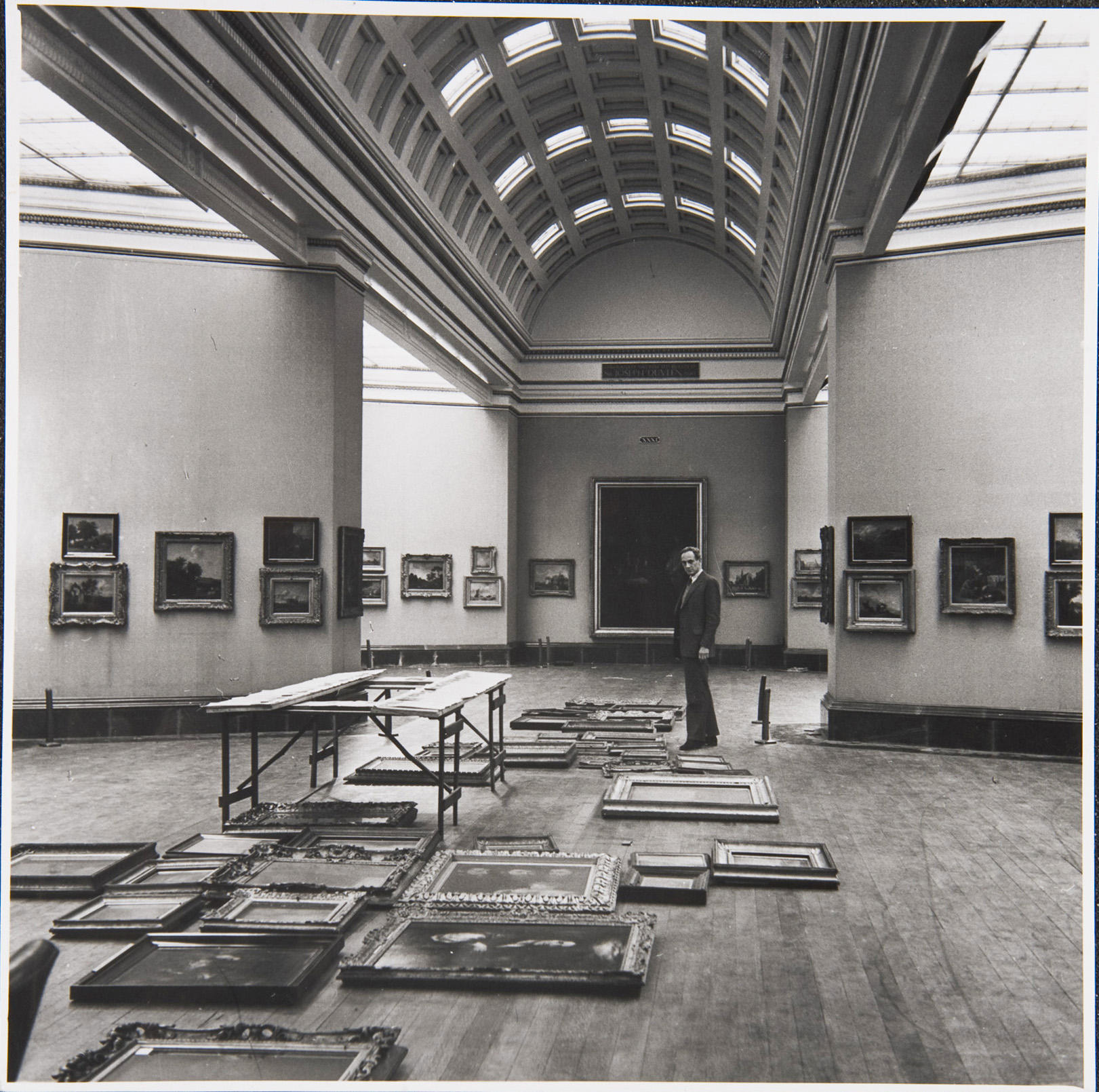 Sir Philip Hendy, Director, standing in Room 29. He is surrounded by paintings laid out on the floor in preparation for a rehang of the room.