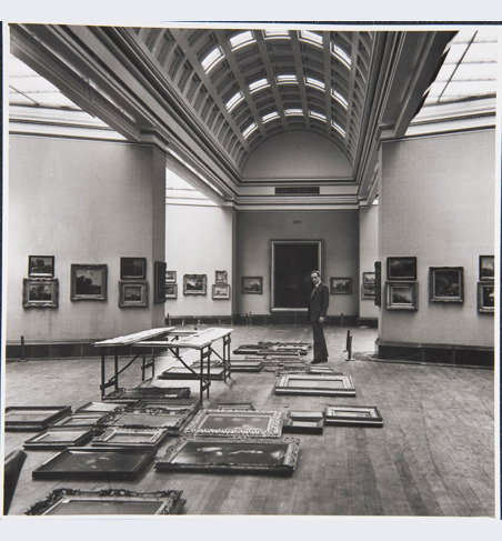 Sir Philip Hendy, Director, standing in Room 29. He is surrounded by paintings laid out on the floor in preparation for a rehang of the room.