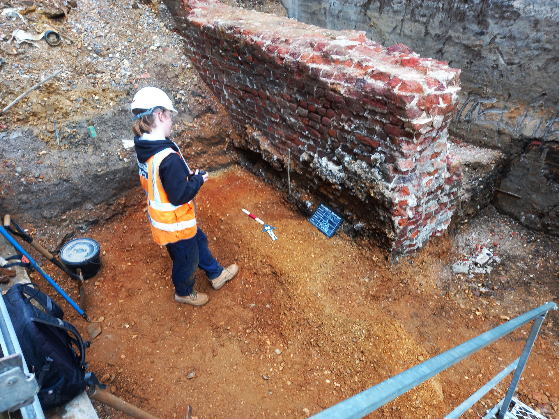 An archaeologist cocks their head in advance of taking a photo of a large red brick wall that has been excavated. The photographed area includes a blackboard with information about the context being photographed, an arrow pointing north, and a red and white metre scale bar.