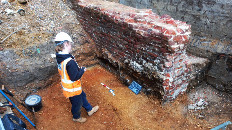 An archaeologist cocks their head in advance of taking a photo of a large red brick wall that has been excavated. The photographed area includes a blackboard with information about the context being photographed, an arrow pointing north, and a red and white metre scale bar.