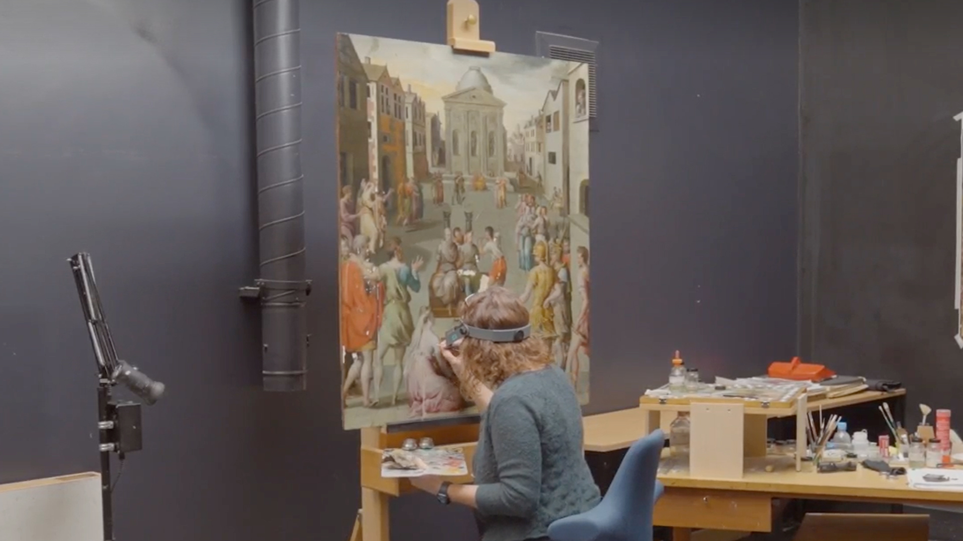 A conservator sits close to a large panel painting on an easel. She is wearing magnifying glasses and holds a fine paintbrush and palette
