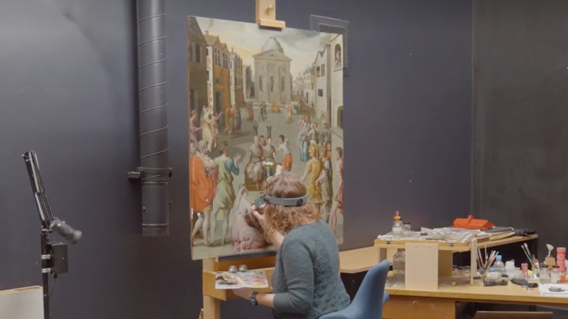 A conservator sits close to a large panel painting on an easel. She is wearing magnifying glasses and holds a fine paintbrush and palette